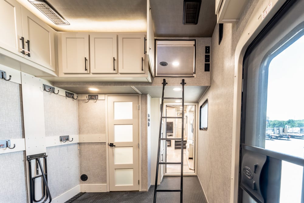 Bright interior of a modern trailer/RV with white upper cabinets, wall hooks, and a metal ladder in a narrow hallway leading to a door and windows to the right