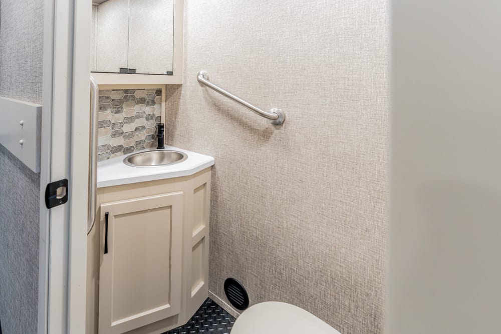 Compact beige bathroom featuring a small vanity with round sink, hex tile backsplash, and wall grab bar. A toilet is in the foreground.