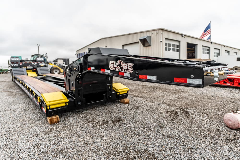 Heavy-duty low-boy trailer with a long gooseneck and Globe Trailers logo, parked on a gravel lot in front of a workshop building with an American flag nearby.
