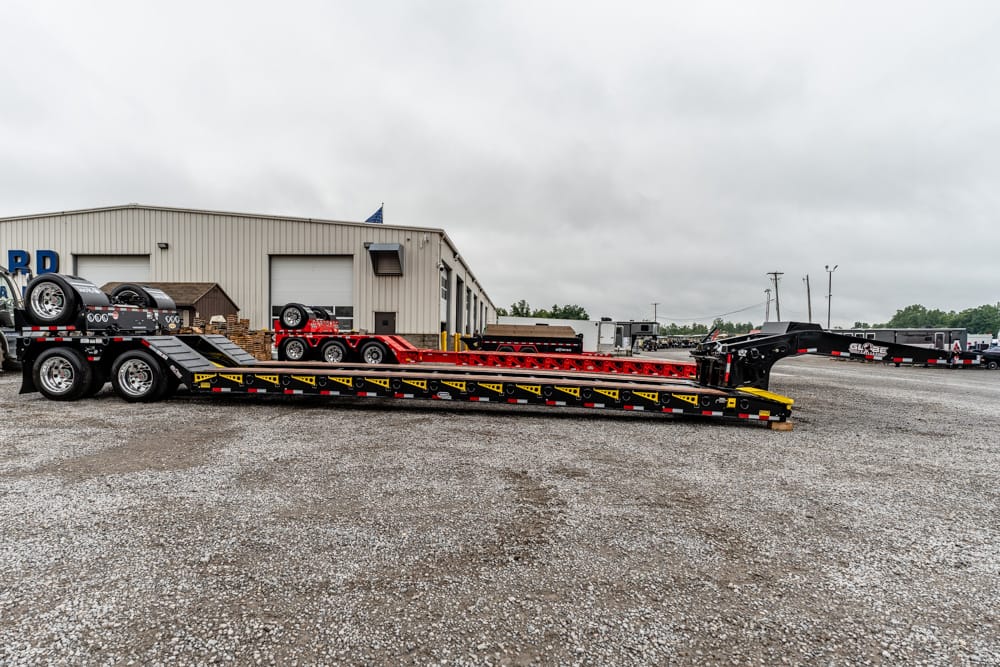Long car hauler trailer with red ramps parked in a gravel lot beside a beige warehouse under an overcast sky, spare tires visible on the trailer's upper deck.