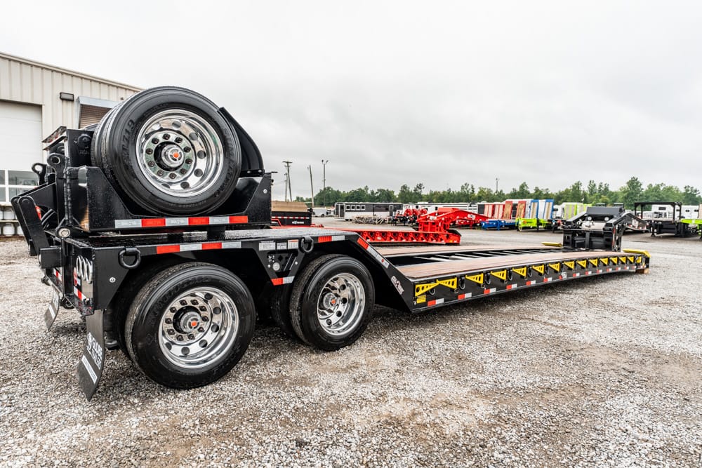 Black heavy‑duty lowboy trailer with a mounted spare wheel at the front, parked on a gravel lot with other equipment in the background.