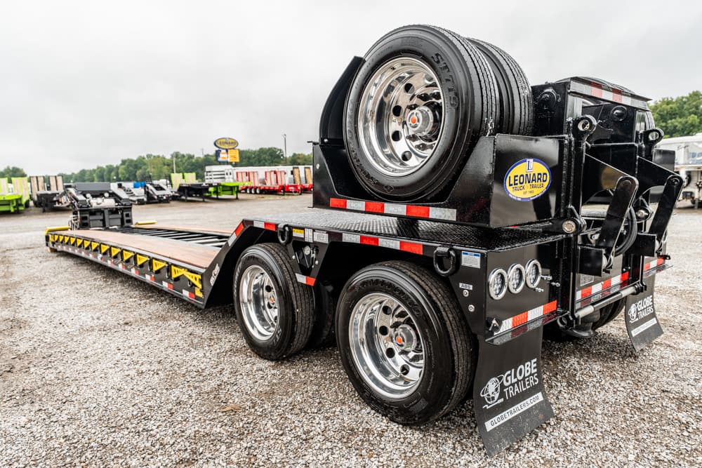 Black heavy-duty flatbed trailer with a mounted spare tire rack and chrome wheels on a gravel lot.