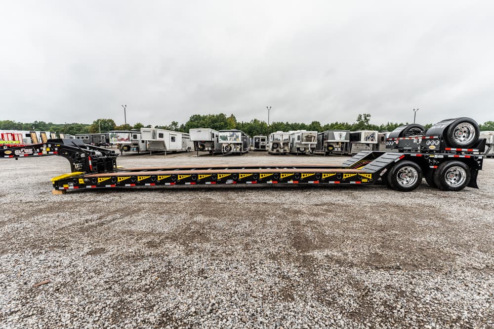 Lowboy trailer with a ramp in a gravel lot, surrounded by various trailers and campers in the background