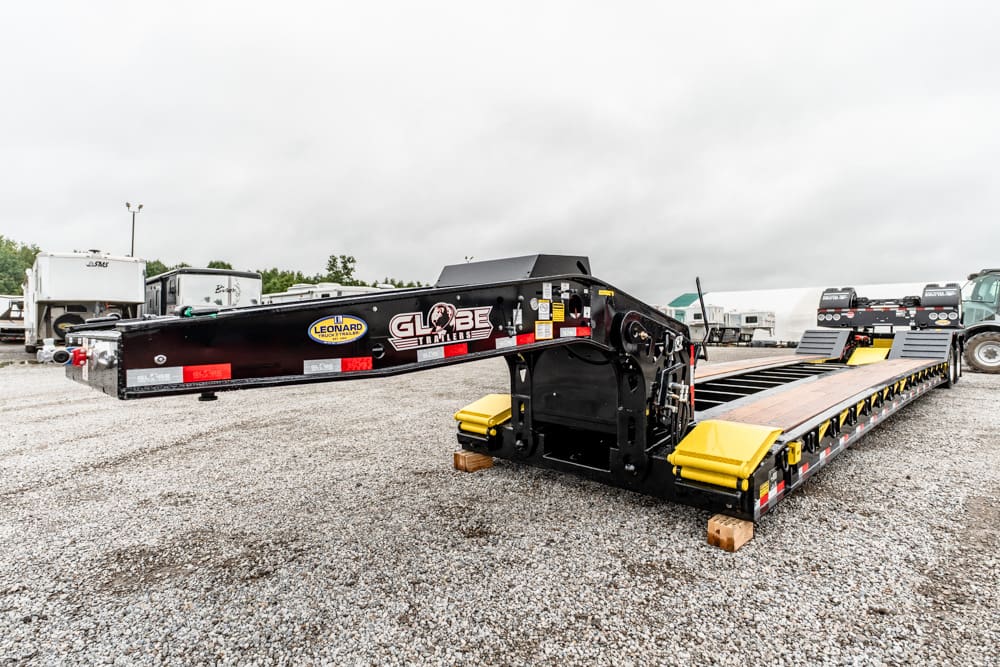 Heavy-duty lowboy trailer with a long black gooseneck, yellow ramps, and industrial equipment in a truck yard.