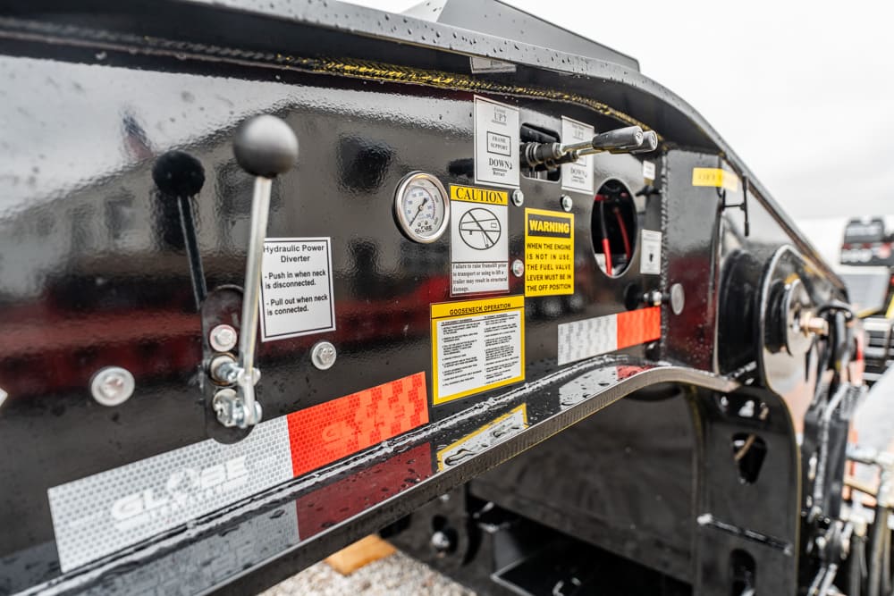 Close-up of a black industrial trailer control panel with levers, gauges, and multiple warning labels stuck to the surface.