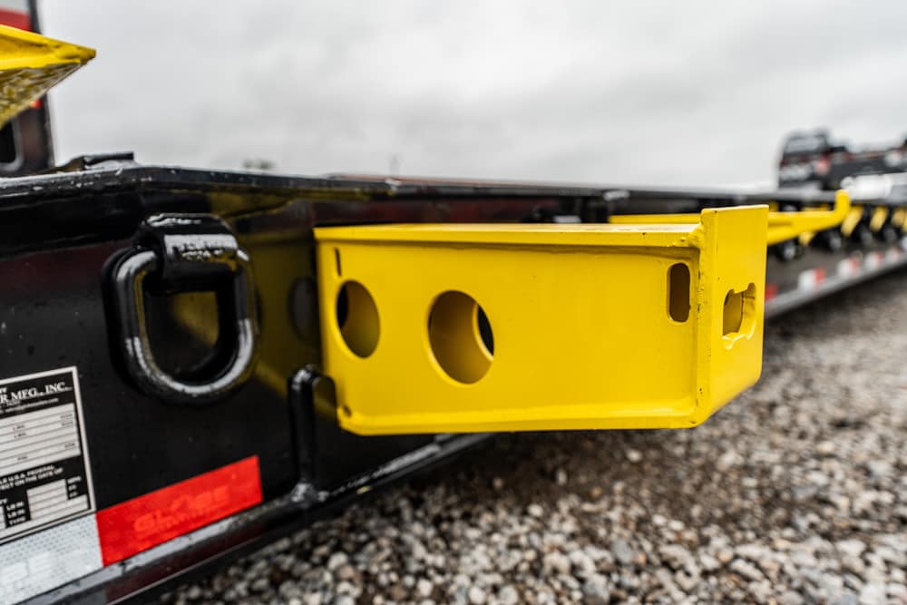 Yellow metal mounting bracket with circular holes attached to a black trailer frame on gravel ground (close-up).