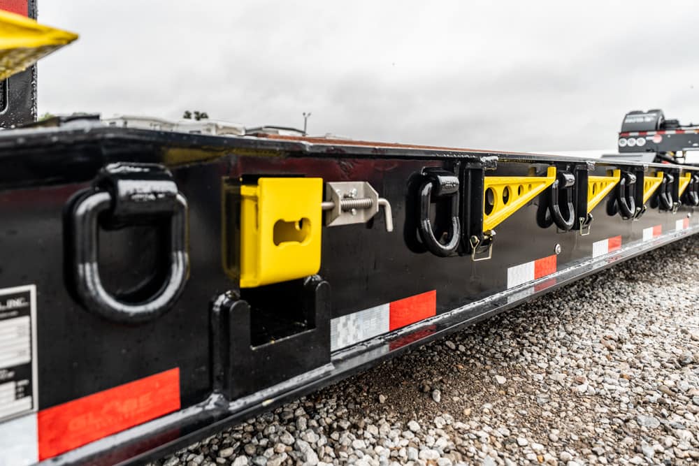 Close-up of a black flatbed trailer side with yellow tie-down brackets and multiple hooks on gravel ground.