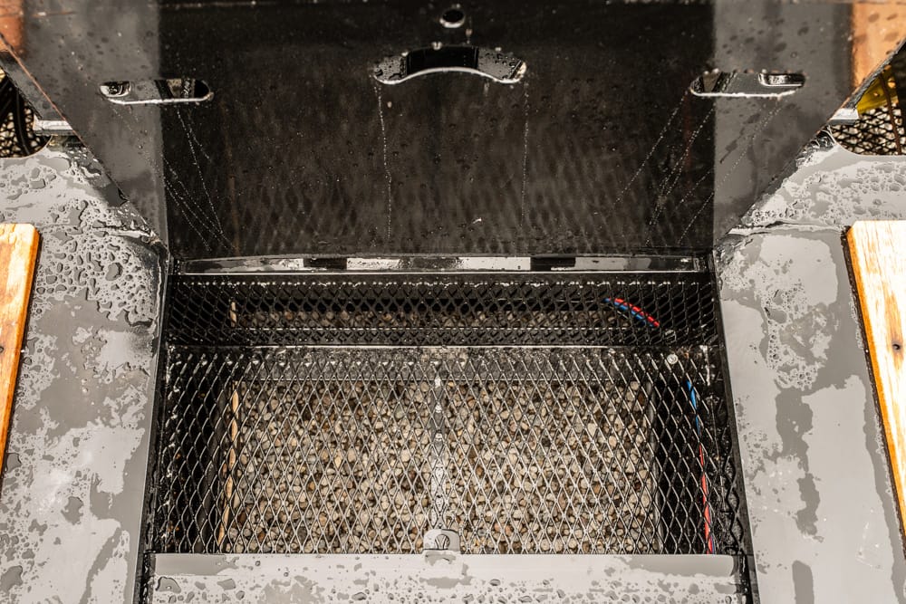 Interior of a charcoal grill with a diamond mesh grate and ash below, opened lid showing handles at the top edge (wet surface around).