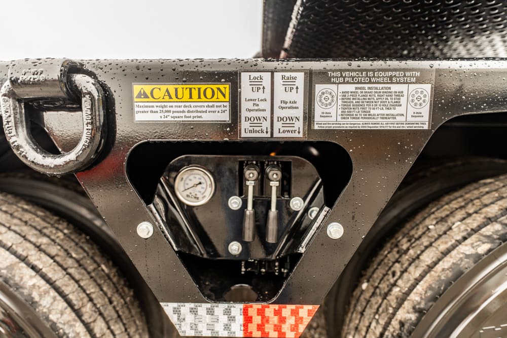Close-up of a trailer hitch coupling with warning labels and a pressure gauge, showing control levers and bolts.