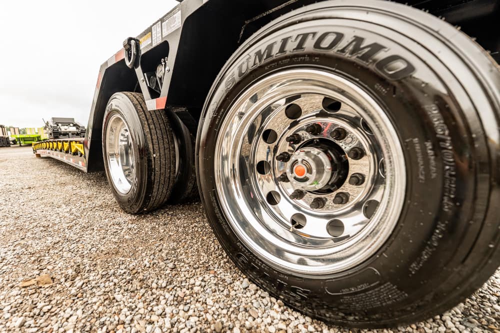 Close-up of a chrome trailer wheel and tire on a gravel lot, with another trailer in the distance in the background