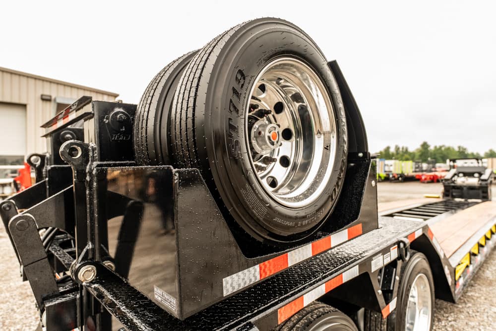 Spare tire mounted on a black flatbed trailer in a gravel lot.