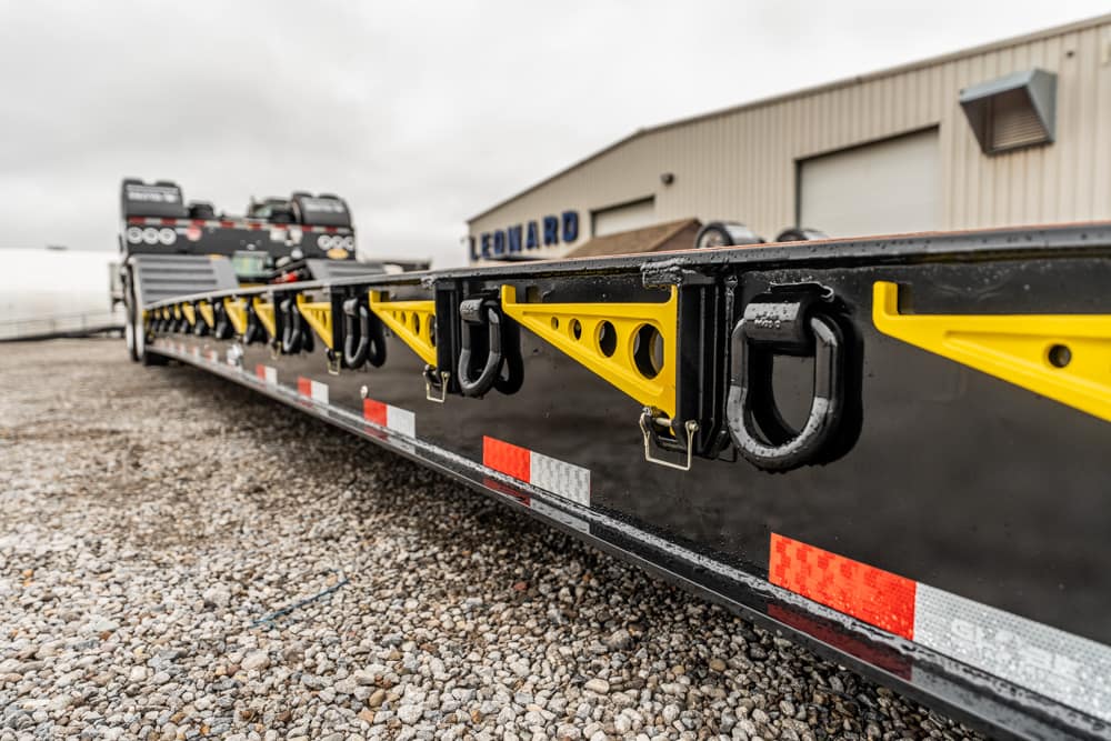 Close-up of a black flatbed trailer with yellow tie-down brackets and metal hooks on a gravel lot, warehouse in the background.