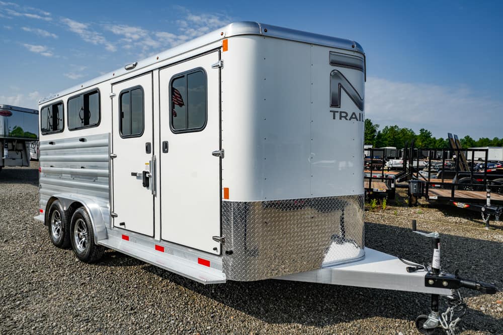 White enclosed cargo trailer with dual axles and diamond-plate front parked on a gravel lot, with other trailers in the background.