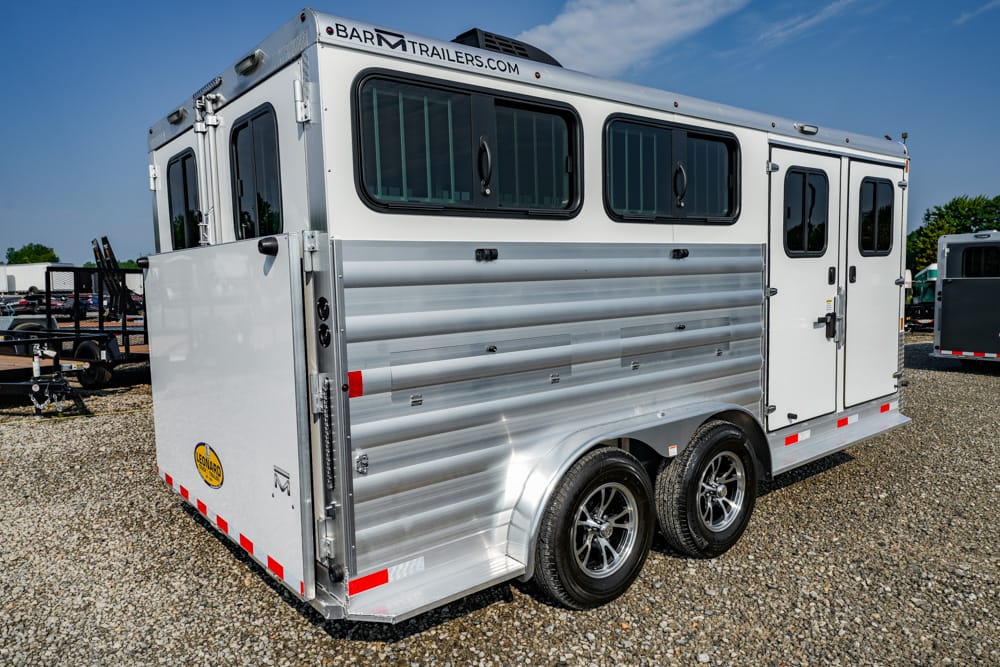 Two-axle aluminum horse trailer with black-framed side windows and a side entry door, parked on a gravel lot under a blue sky.