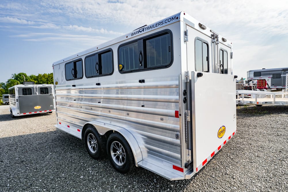 Silver aluminum livestock trailer with tandem axles and three horizontal windows, parked on a gravel lot with other trailers in the background, and BarnTrailers.com branding on top edge.