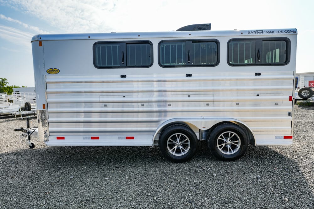 Silver horse trailer with three side windows and dual-axle wheels on a gravel lot