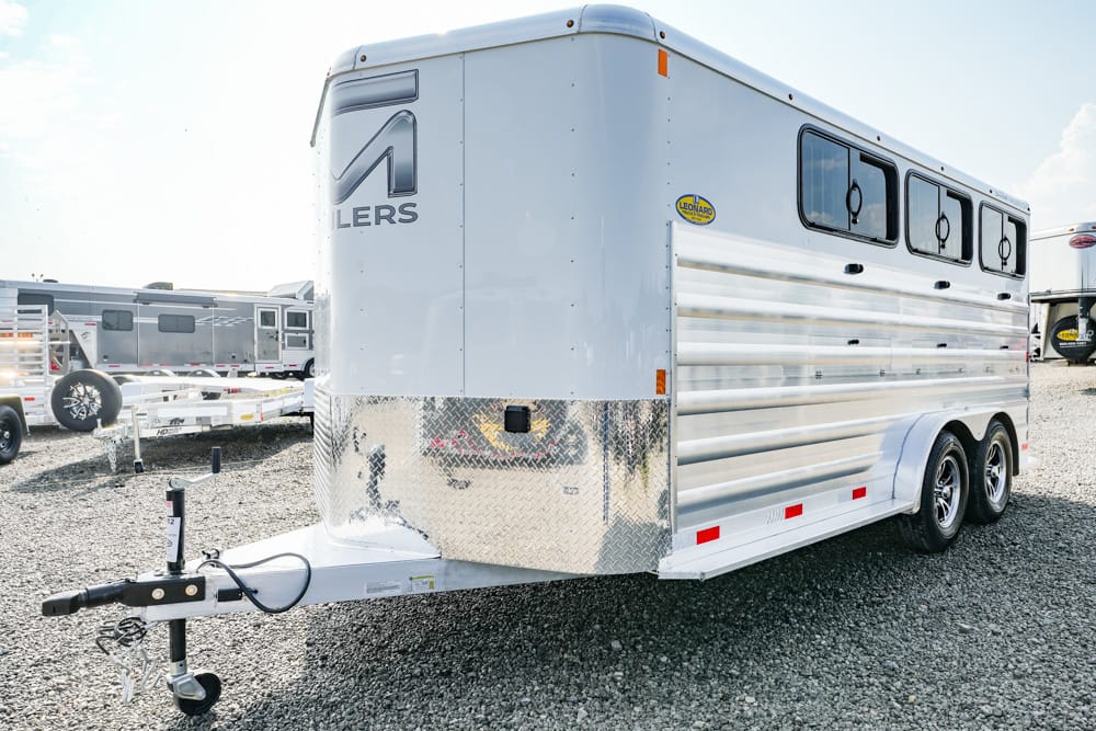 White horse trailer with diamond-plate front and dual axles parked on a gravel lot, hitch visible in foreground.