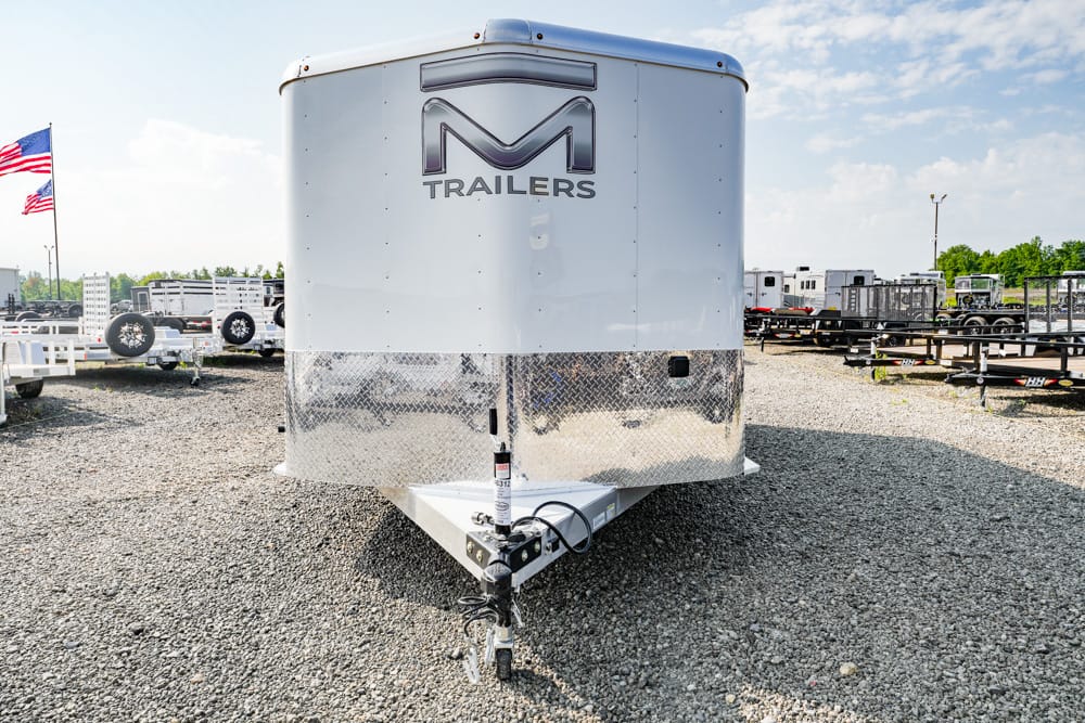 Front view of a white M Trailers enclosed trailer with a diamond-plate lower panel and hitch, parked on a gravel lot at a trailer lot.