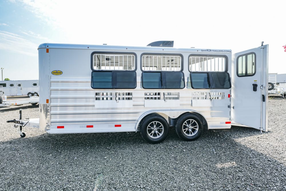 Silver horse trailer with open rear door and dual axles on a gravel lot, windows along the side and protection bars over stalls