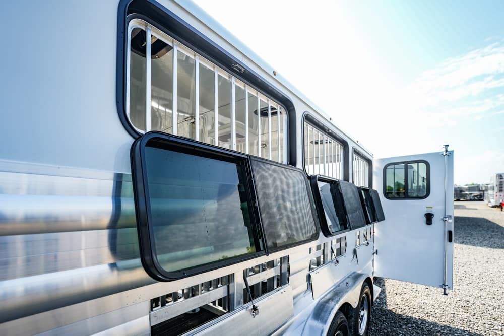 White horse trailer with ventilated side windows and an open rear door on a gravel lot original sunlight casting reflections on the metal surface.
