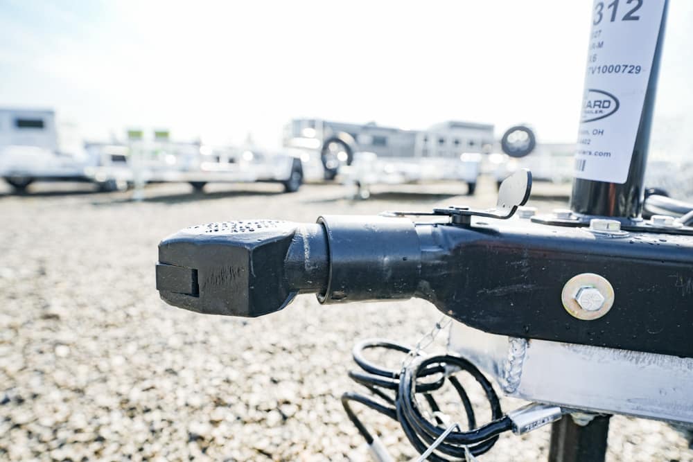 Close-up of a black trailer hitch on a boat trailer, with a tag and coiled wires in the foreground, a gravel lot and trailers in the background.