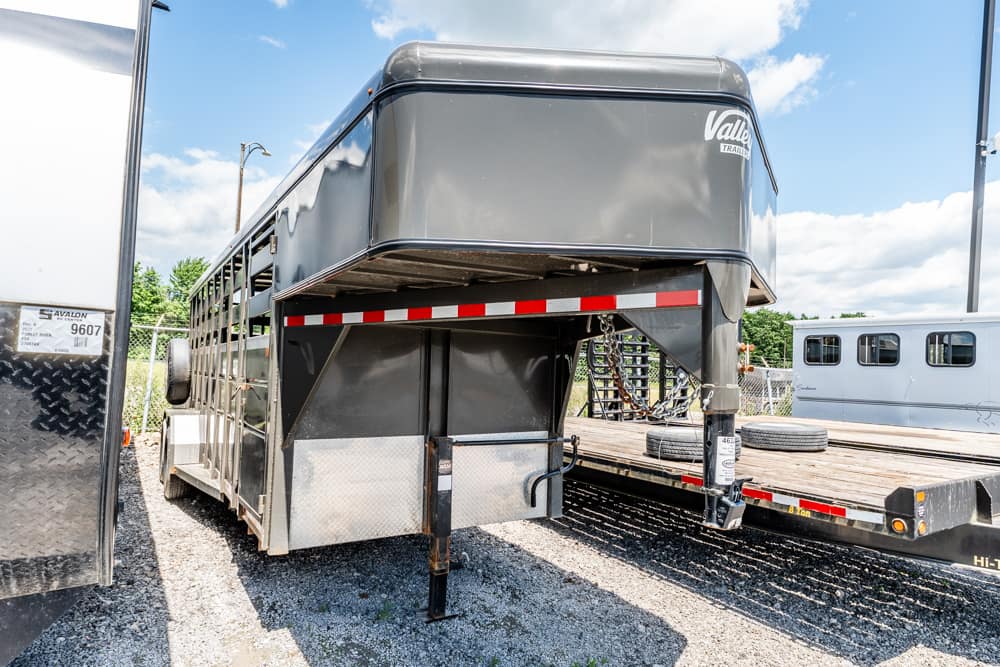 Gooseneck horse trailer in a gravel lot, raised hitch and safety chains, with a flatbed trailer nearby.