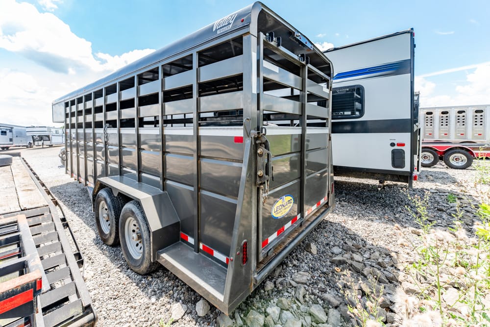 Rear and side view of a dark gray livestock trailer with slatted sides on a gravel lot, another trailer visible in the background.