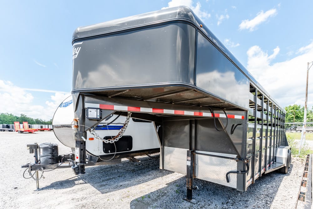 Livestock trailer with black metal body and slatted side panels parked on gravel, hitch and safety chains visible in front.