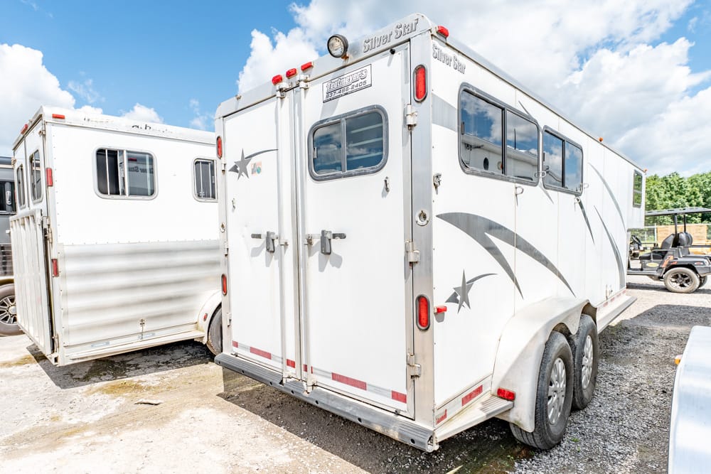 White two-horse trailer with dual axles, side windows, and rear double doors on a gravel lot.