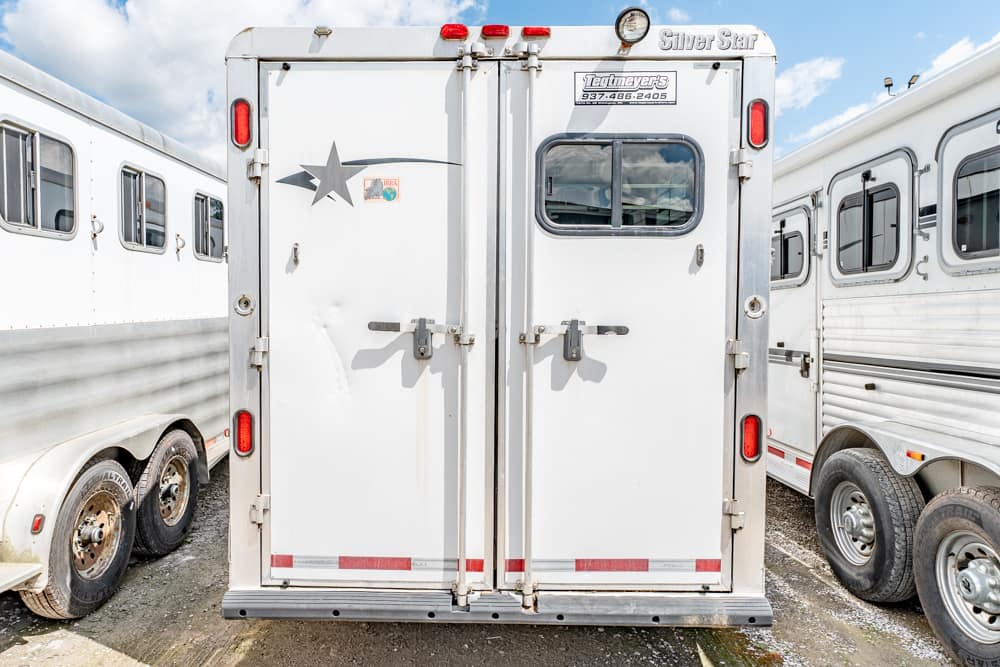 White double-horse trailer rear with two metal doors, small window, red lights, and adjacent trailers parked on a dirt lot.