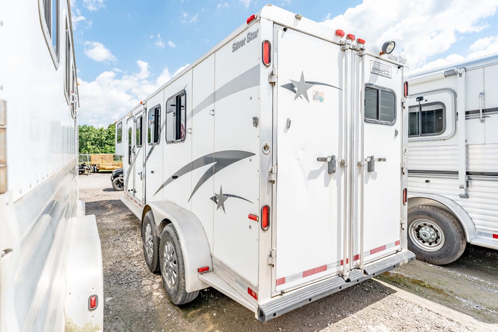 White horse trailer with star decals parked among other trailers on a gravel lot under a sunny sky