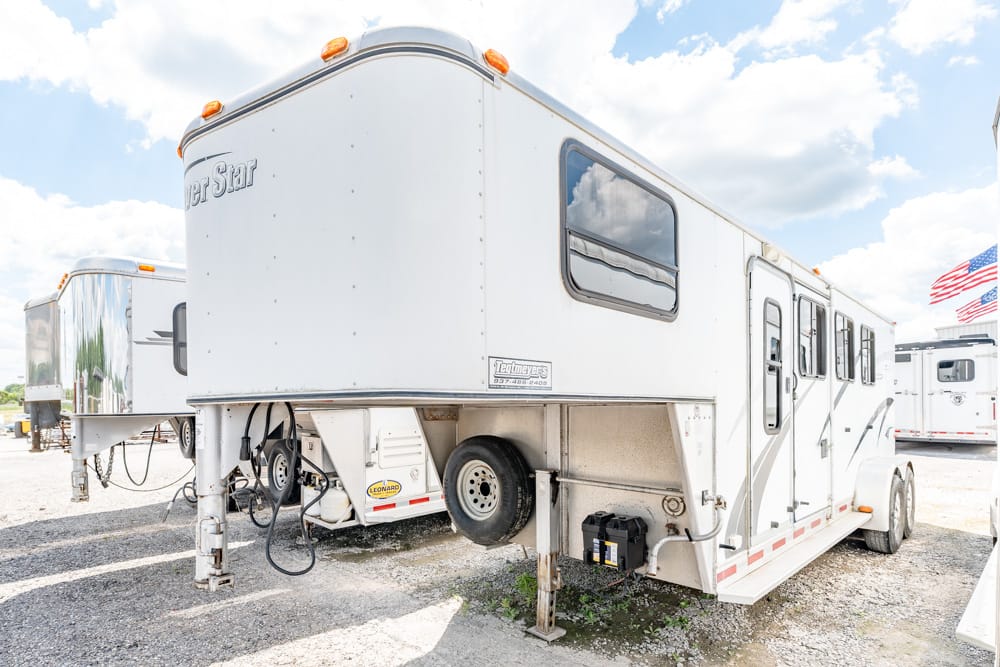 White gooseneck fifth-wheel RV trailer parked in a gravel lot with other trailers nearby, front hitch visible and spare tire attached undercarriage.