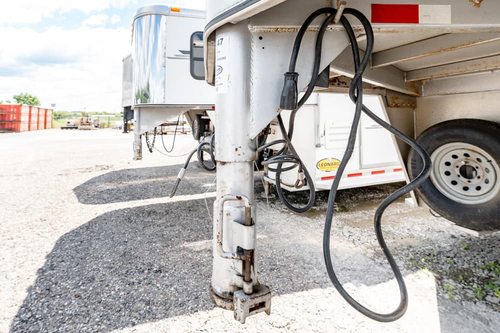 Close-up of a rusted trailer landing gear and tangled hoses under a parked semi-trailer on a gravel lot.