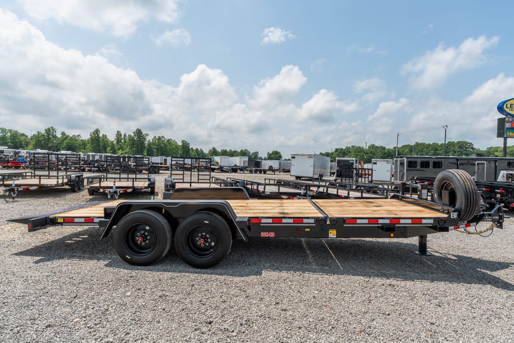 Flatbed trailer with a wooden deck and dual rear axles sits on a gravel lot at a trailer dealership, with a spare tire mounted at the front and other trailers in the background.