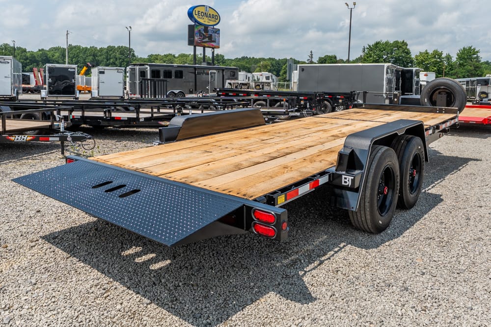Dual-axle flatbed trailer with a wooden deck and a fold-down loading ramp on a gravel lot