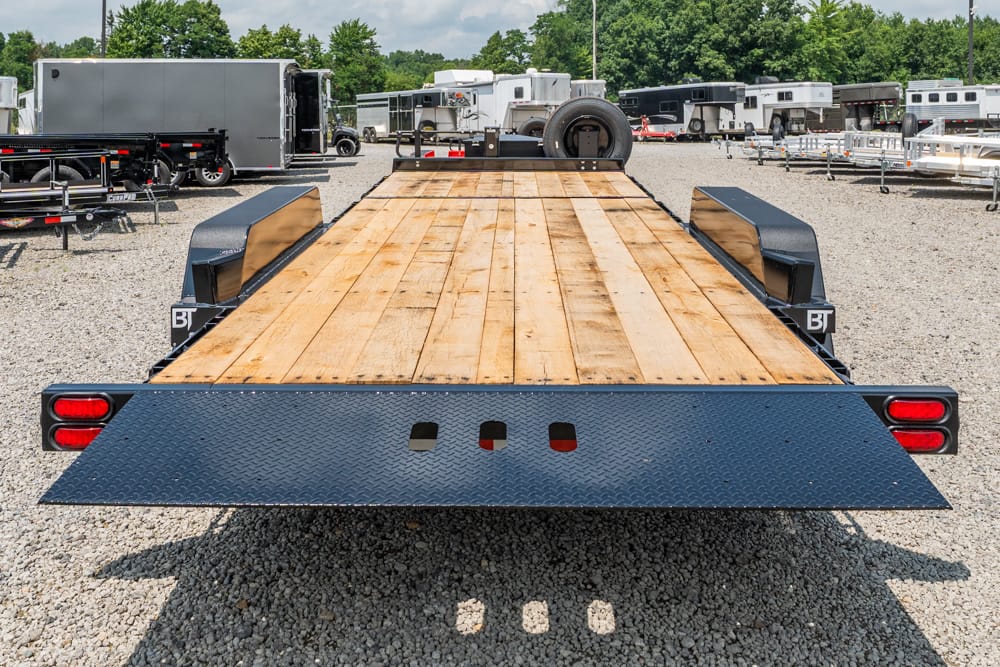 Low-angle view of a flatbed trailer with a wooden deck parked in a gravel trailer yard, red taillights visible on the rear.