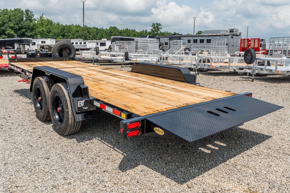 Dual-axle flatbed trailer with a wooden deck and rear ramp on a gravel lot.