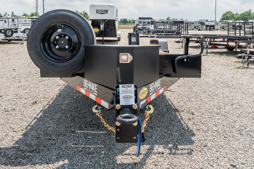 Front view of a black equipment trailer with a spare tire on the left and a hitch, in a gravel lot with other trailers nearby.