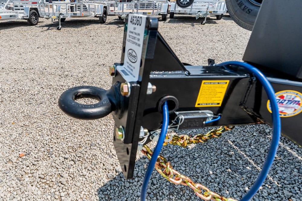 Trailer hitch assembly with safety chain, blue electrical cable, and ring shackle on a gravel lot with trailers in background (informative)