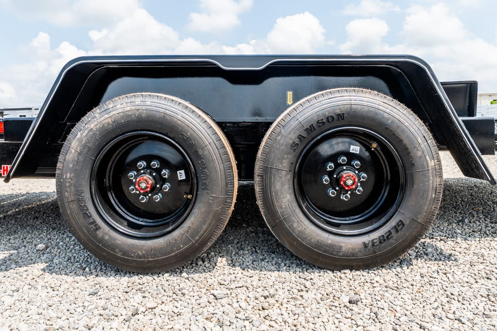Dual trailer wheels with black rims and red hub nuts on a gravel lot under a blue sky with white clouds
