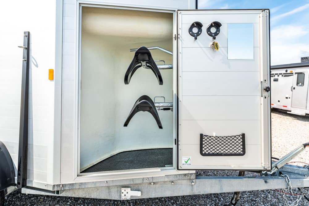 Interior of a horse trailer with two saddles hanging on racks inside, doorway open to the left side of the image.