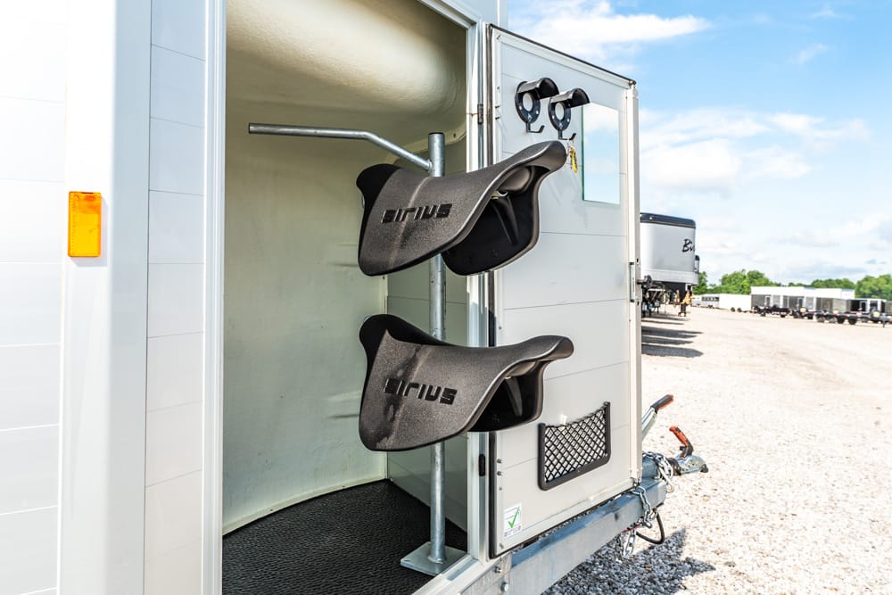 Two black saddles hanging on a rack inside an open horse trailer, with a gravel lot and other trailers in the background