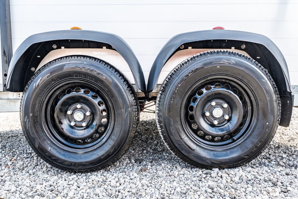 Twin trailer wheels with black steel rims and fenders on a gravel surface under a white wall.