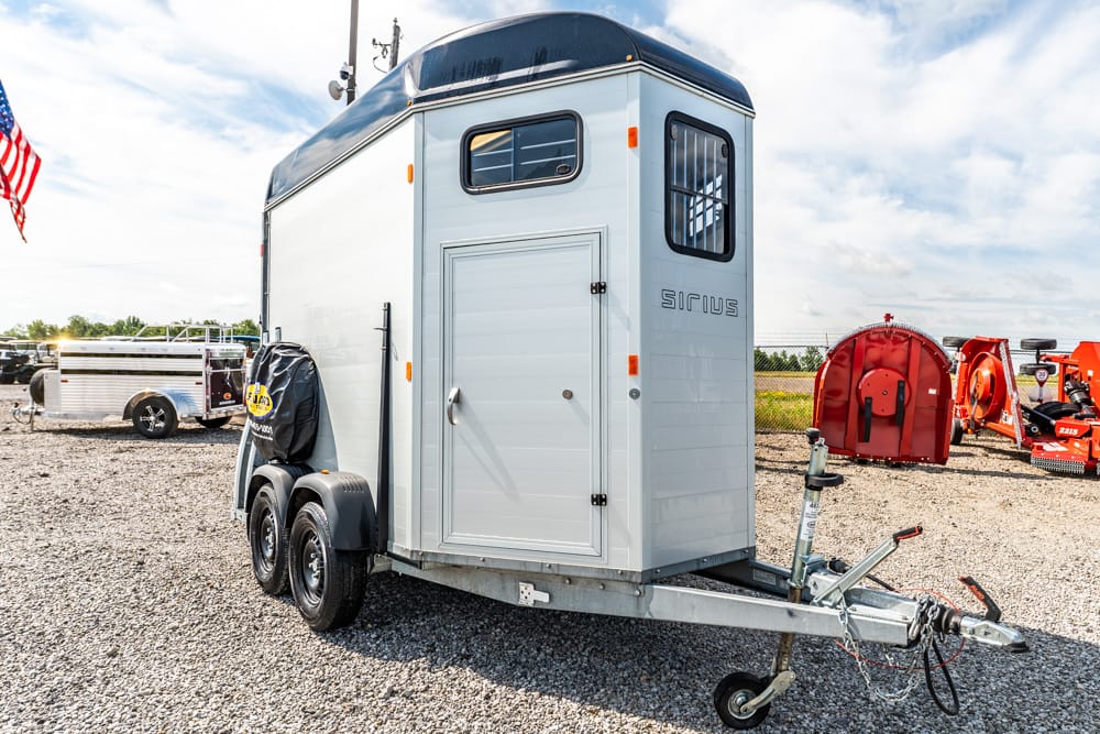 Gray enclosed horse trailer on a dual-axle trailer, hitched to a tow bar in a gravel lot with farm equipment nearby.