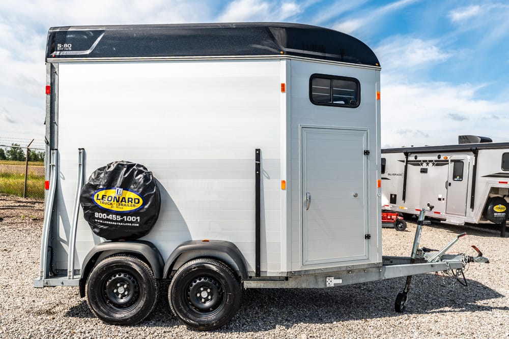 Silver horse trailer with dual axles and a spare tire cover reading 'Leonard Truck & Trailers' parked on gravel with a front hitch and side door closed under a blue sky.