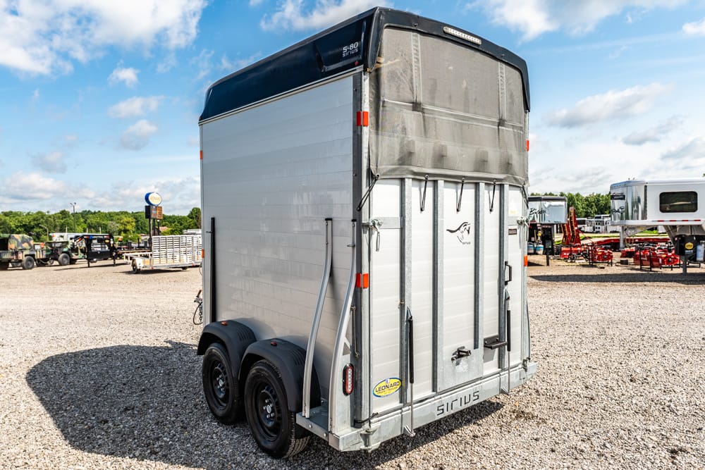 Rear view of a silver, two-axle horse trailer with a black top parked on a gravel lot; other trailers visible in the background.