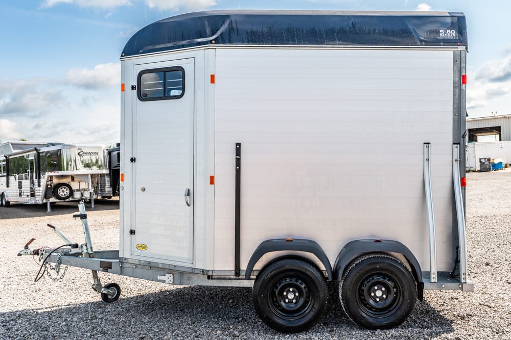 White horse trailer with dual axles and a front hitch parked on a gravel lot, with other trailers in the background.