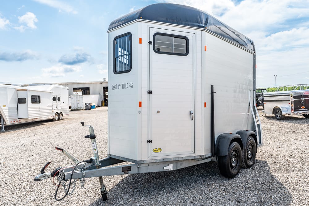 White enclosed horse trailer on a dual-axle trailer, parked on a gravel lot with other trailers in the background under a blue sky