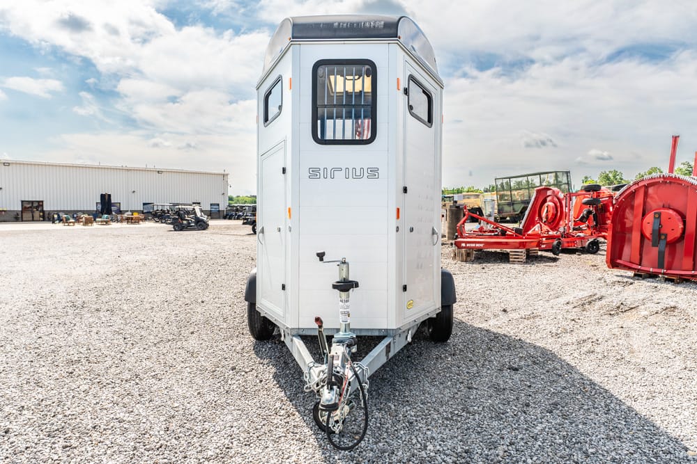 White horse trailer on a gravel lot with red farm machinery in the background and a blue sky above.