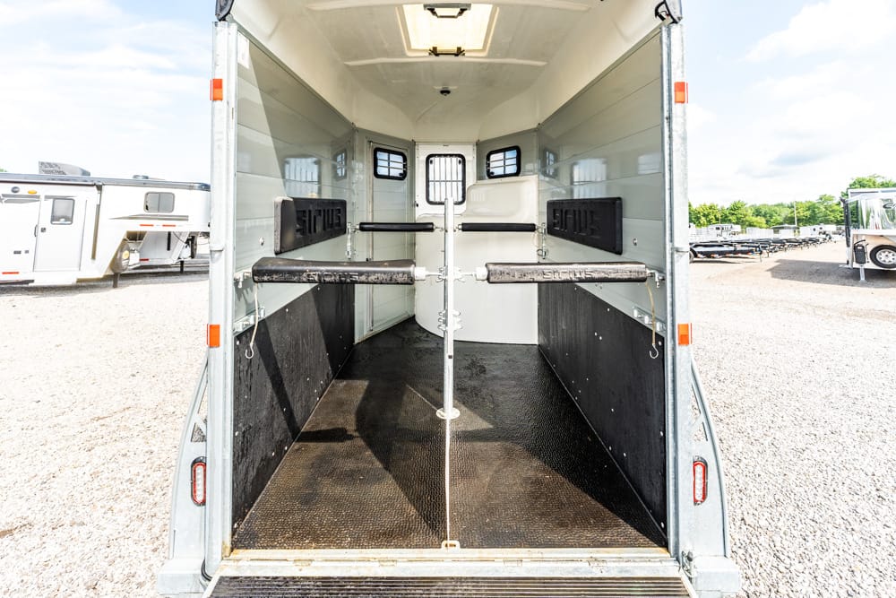Interior of a two-stall horse trailer with divider rails and padded lower walls, viewed from the open rear doors on a gravel lot